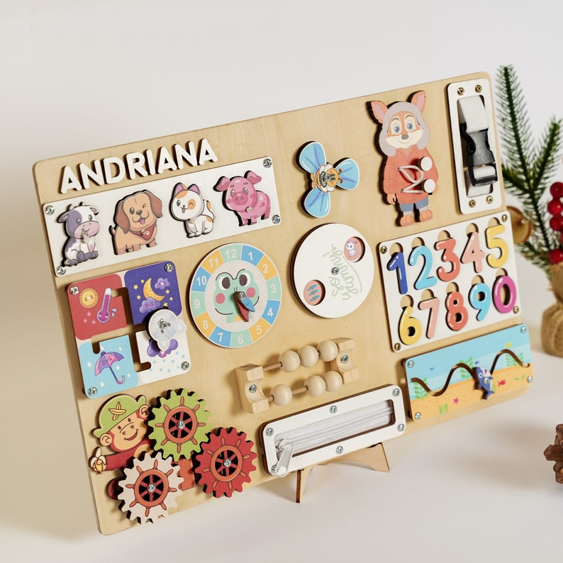 May include: Wooden activity board featuring various interactive elements for children. Includes animal figures, a clock, numbers, gears, a zipper, and a spinner. The name "ANDRIANA" is displayed at the top. The board is designed to promote learning and fine motor skills.