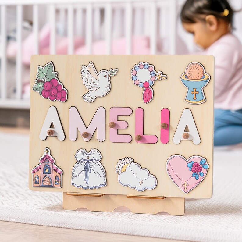 May include: A wooden puzzle board with a pink and white colour scheme. The board features the name "AMELIA" in large, colourful letters. There are also various religious symbols and images, including a dove, a church, a heart, and a rosary.