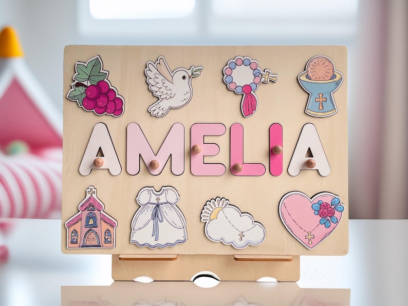 May include: A wooden puzzle board with a pink and white theme. The board features the name "AMELIA" in pink letters and various religious symbols, including a dove, a chalice, a rosary, and a heart with a cross. The board is designed for children to learn their name and religious symbols.