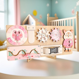 May include: Wooden activity board featuring a pink clock with a frog face, gears, a buckle, and a sliding track with a fish design. A cartoon bunny in a pink coat is also included. The board is designed for children.