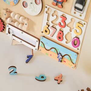 May include: Wooden activity board featuring various interactive elements. Includes a zipper, abacus, and a number puzzle with colorful, removable numbers. A small beach scene with a shark is also present. The board is designed for early learning and sensory exploration.