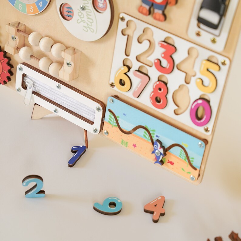 May include: Wooden activity board featuring various interactive elements. Includes a zipper, abacus, and a number puzzle with colorful, removable numbers. A small beach scene with a shark is also present. The board is designed for early learning and sensory exploration.