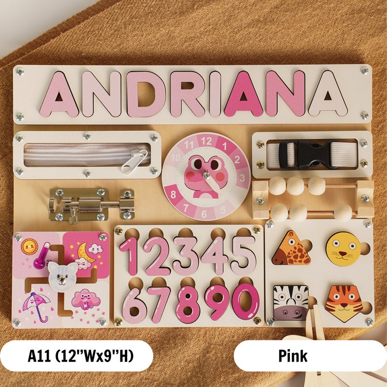 May include: A pink wooden busy board with the name "ANDRIANA" at the top. The board has a variety of activities for children, including a zipper, a latch, a clock, a bead maze, a number puzzle, and a shape puzzle. The board is 12 inches wide and 9 inches high.