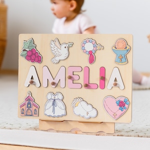 May include: A wooden puzzle board with the name Amelia spelled out in pink letters. The board features various religious symbols and images, including a dove, a church, a rosary, and a chalice.