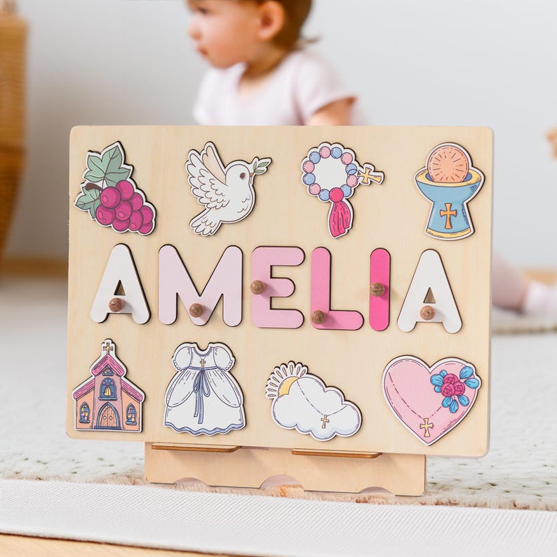 May include: A wooden puzzle board with the name Amelia spelled out in pink letters. The board features various religious symbols and images, including a dove, a church, a rosary, and a chalice.