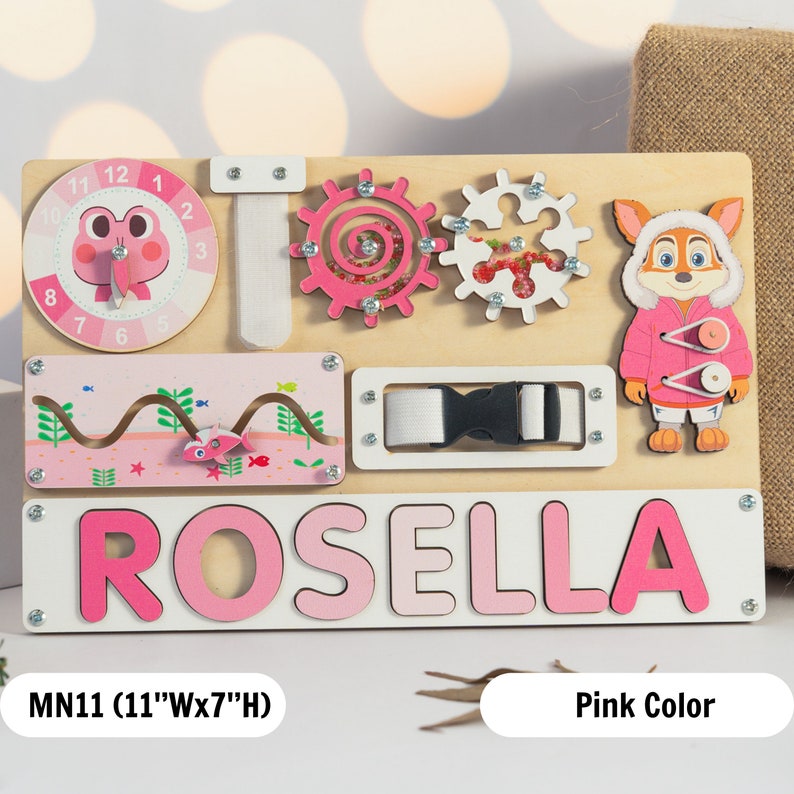 May include: A pink wooden busy board with a clock, a buckle, a maze, two gears, a frog, a fox, and the name "ROSELLA" spelled out in large pink letters. The board measures 11 inches wide by 7 inches high.