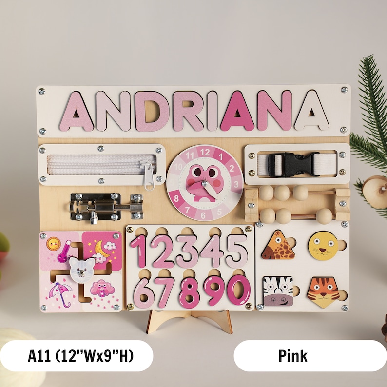 May include: A pink wooden activity board with the name "ANDRIANA" spelled out in pink letters. The board features a variety of activities for children, including a zipper, a latch, a clock, a bead maze, and a number puzzle. The board measures 12 inches wide and 9 inches high.