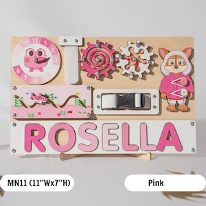 May include: A pink wooden activity board with a clock, gears, a buckle, a zipper, and a name puzzle that says "ROSELLA". The board is 28 cm wide and 18 cm high.