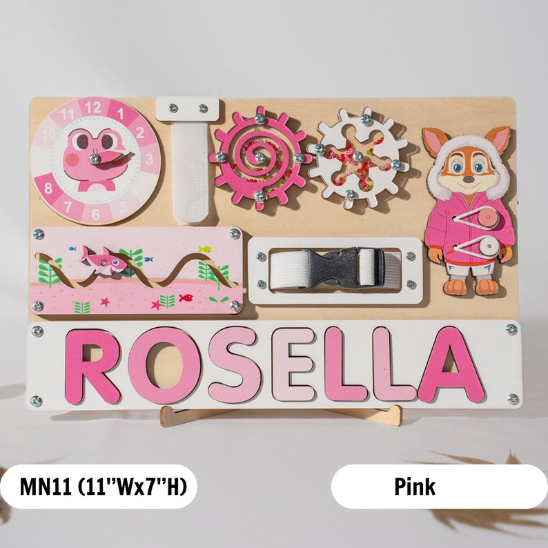 May include: A pink wooden activity board with a clock, gears, a buckle, a zipper, and a name puzzle that says "ROSELLA". The board is 28 cm wide and 18 cm high.