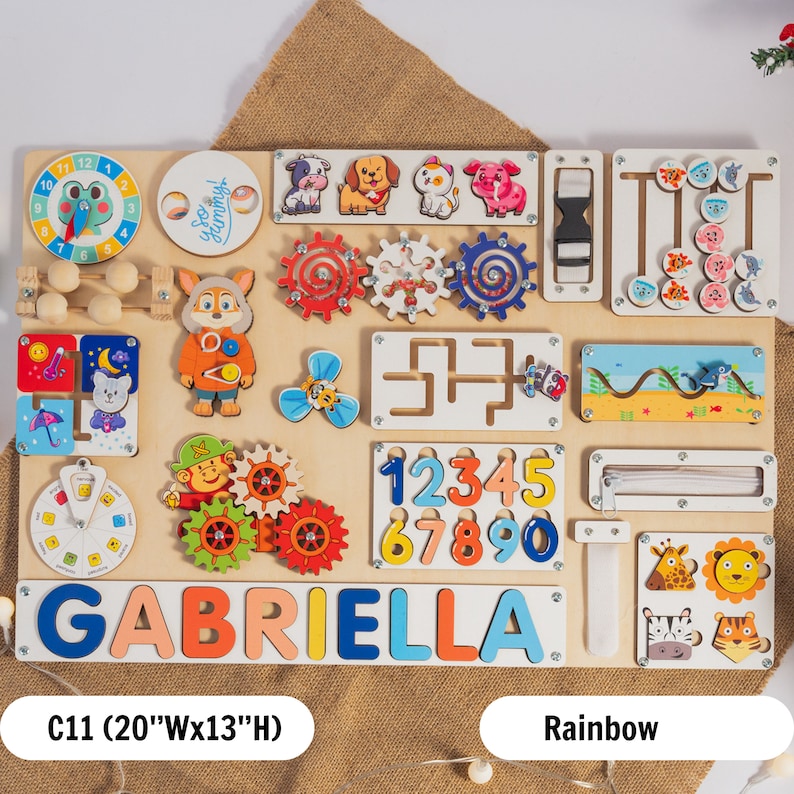 May include: A colorful wooden activity board with various features for children, including a clock, gears, a zipper, a maze, a slide, and a name puzzle that reads "GABRIELLA". The board is 20 inches wide and 13 inches high.