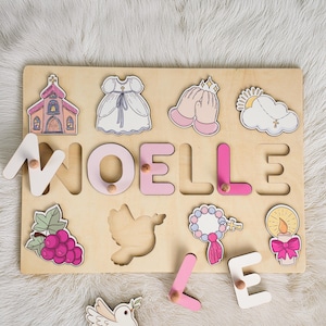May include: A wooden puzzle featuring the name "NOELLE" spelled out in pink and white letters. The puzzle includes images of a church, a dove, a communion dress, a rosary, a candle, and a cloud with a cross.