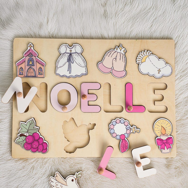 May include: A wooden puzzle featuring the name "NOELLE" spelled out in pink and white letters. The puzzle includes images of a church, a dove, a communion dress, a rosary, a candle, and a cloud with a cross.