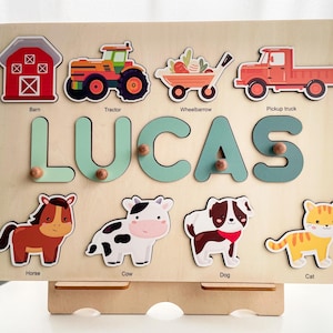 May include: A wooden educational toy with a name puzzle that says "LUCAS" and images of farm animals, vehicles, and a barn. The toy is on a stand.