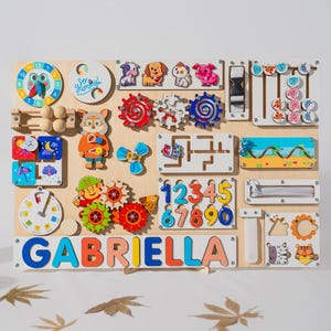 May include: A wooden activity board with various colorful shapes, gears, and numbers. The board has a clock, a maze, a zipper, and a sliding door. The name "GABRIELLA" is spelled out in colorful letters at the bottom.