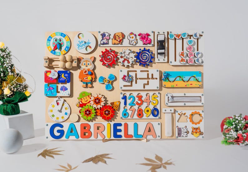 May include: A wooden activity board with various colorful shapes, gears, and numbers. The board has a clock, a maze, a zipper, and a sliding door. The name "GABRIELLA" is spelled out in colorful letters at the bottom.