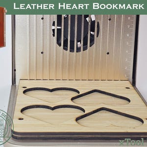 May include: A laser engraving machine with a wooden jig for making heart-shaped leather bookmarks. The machine has a metal frame, a fan, and a ruler. The text "LEATHER HEART BOOKMARK JIG" is visible.