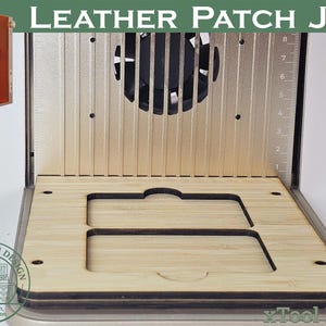 Op de afbeelding: Een lederen patchmal met de tekst "LEATHER PATCH JIG" bovenaan. De mal heeft een houten basis met uitsparingen, een metalen achterkant met een ventilator en een liniaal. Een oranje XTOOL-machine staat in de linkerbovenhoek.
