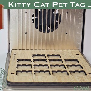 May include: A laser engraving machine with the text "KITTY CAT PET TAG JIG". The machine has a wooden base with cat-shaped cutouts. An orange laser module is on the left. The machine is designed for pet tag creation.