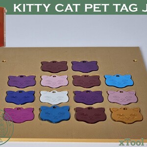 May include: A gold-coloured rectangular jig plate featuring twelve recessed cat-shaped slots, each containing a cat-shaped pet tag in various colours including blue, purple, pink, white, and gold. The text KITTY CAT PET TAG JIG appears at the top.