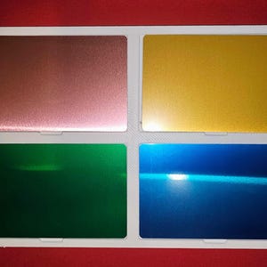 May include: A white tray containing four rectangular metallic colour samples: copper, gold, green, and blue. Each sample has a reflective, textured surface. The background is a solid red.