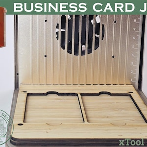 May include: A business card jig with the text "BUSINESS CARD JIG" at the top. The jig has a wooden base with two card slots, a metal back with a fan, and a ruler on the side. An orange laser cutter is in the upper left corner.