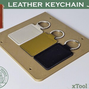 Puede incluir: Una plantilla para llaveros de cuero con tres llaveros en blanco, verde oliva y negro. Cada llavero tiene un anillo plateado. La plantilla es de color beige y tiene el texto "LEATHER KEYCHAIN JIG". Una caja naranja está en la esquina superior izquierda.