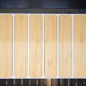 May include: Six rectangular bamboo bookmarks with rounded corners and a hole at the top for a string. The bookmarks are light brown with a natural wood grain and are set in a white frame.