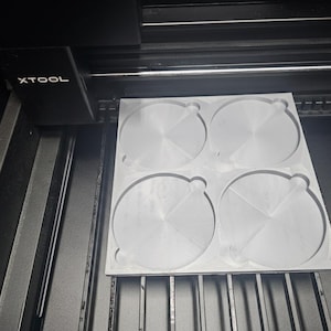 May include: A laser cutter with a gray, circular design cut into a rectangular piece of material. The machine is black with the brand name "XTOOL" visible on the side.