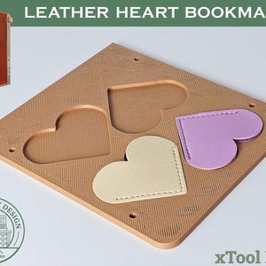 Pode incluir: Um conjunto de marcadores de livros em couro em forma de coração bege com um design cortado a laser. O conjunto inclui uma base bege com recortes em forma de coração e três marcadores em forma de coração em creme e lavanda.