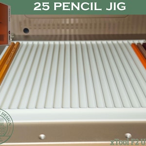 May include: A white pencil jig with the text "25 PENCIL JIG" and "xTool F2 Ultra UV". Several pencils in various colors are placed in the jig. An orange and brown box is in the upper left corner.