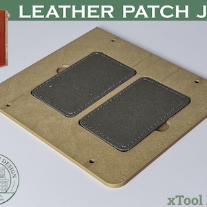 Puede incluir: Una plantilla de parche de cuero beige con dos parches de cuero gris. La plantilla tiene forma cuadrada con esquinas redondeadas y varios agujeros pequeños. El texto "LEATHER PATCH JIG" está en la parte superior. La imagen también incluye una pequeña máquina naranja y un logotipo.