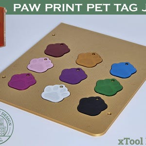 May include: A gold-coloured rectangular jig with a textured surface featuring nine paw-print-shaped pet tags in various colours including red, pink, purple, blue, green, black, and white, designed for use with the xTool F1/F2 laser machine.