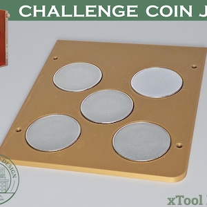 Puede incluir: Una plantilla para monedas de color bronceado con cinco huecos circulares, cada uno con una moneda de plata. El texto "CHALLENGE COIN JIG" está en la parte superior. Una máquina naranja está en la esquina superior izquierda. El texto "xTool F1/F2" está en la parte inferior.
