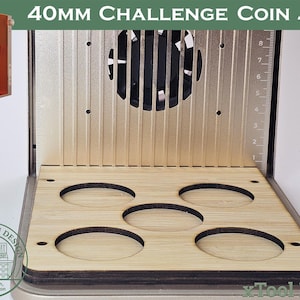 Puede incluir: Una plantilla para monedas con el texto "40MM CHALLENGE COIN JIG". La plantilla tiene una base de madera con cinco recortes circulares. Detrás de la base hay una estructura metálica con un ventilador y una regla. A la izquierda hay una caja naranja con un asa.