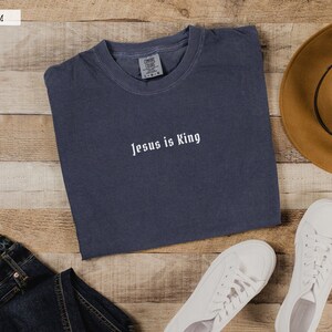 May include: A dark blue t-shirt with the text "Jesus is King" in white lettering. The t-shirt is laid flat on a wooden surface with a pair of white sneakers, a pair of blue jeans, and a brown cowboy hat.