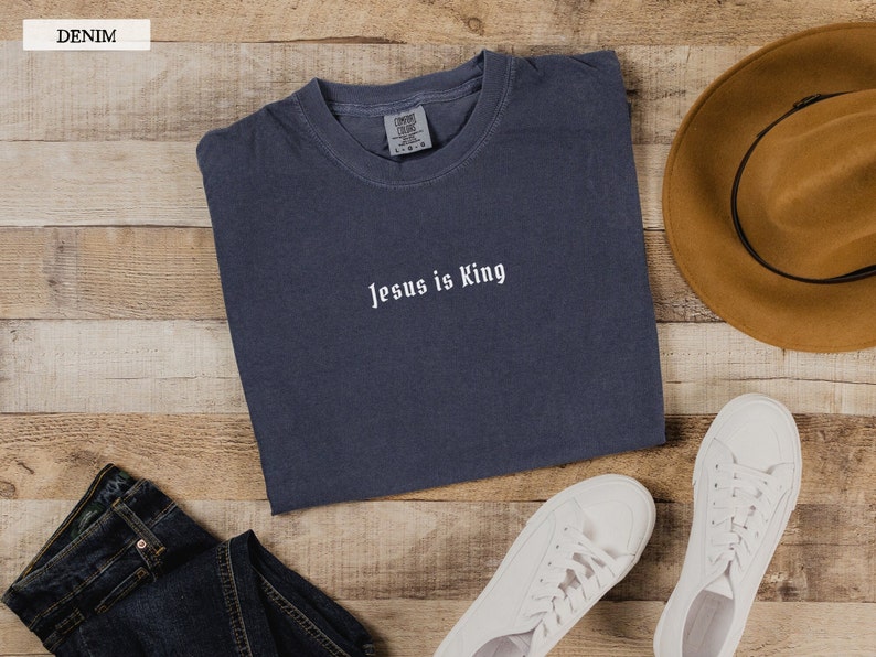 May include: A dark blue t-shirt with the text "Jesus is King" in white lettering. The t-shirt is laid flat on a wooden surface with a pair of white sneakers, a pair of blue jeans, and a brown cowboy hat.