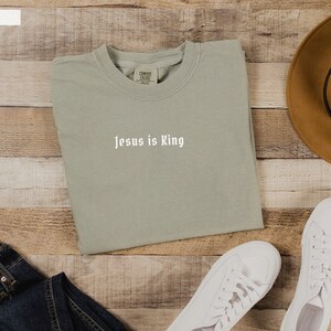 May include: A light green t-shirt with the text "Jesus is King" in white lettering. The t-shirt is folded and laid on a wooden surface with a pair of white sneakers, a pair of blue jeans, and a brown hat.