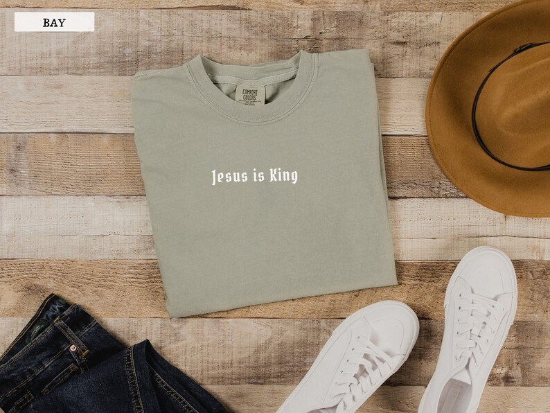 May include: A light green t-shirt with the text "Jesus is King" in white lettering. The t-shirt is folded and laid on a wooden surface with a pair of white sneakers, a pair of blue jeans, and a brown hat.
