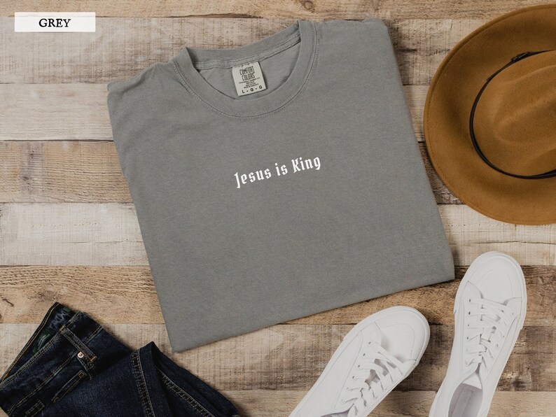 May include: A gray t-shirt with the text "Jesus is King" printed in white on the front. The shirt is laid flat on a wooden surface with a brown hat and white sneakers.