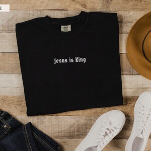 May include: Black long-sleeved t-shirt with the text "Jesus is King" in white lettering. The shirt is laid flat on a wooden surface with a brown hat and white sneakers.