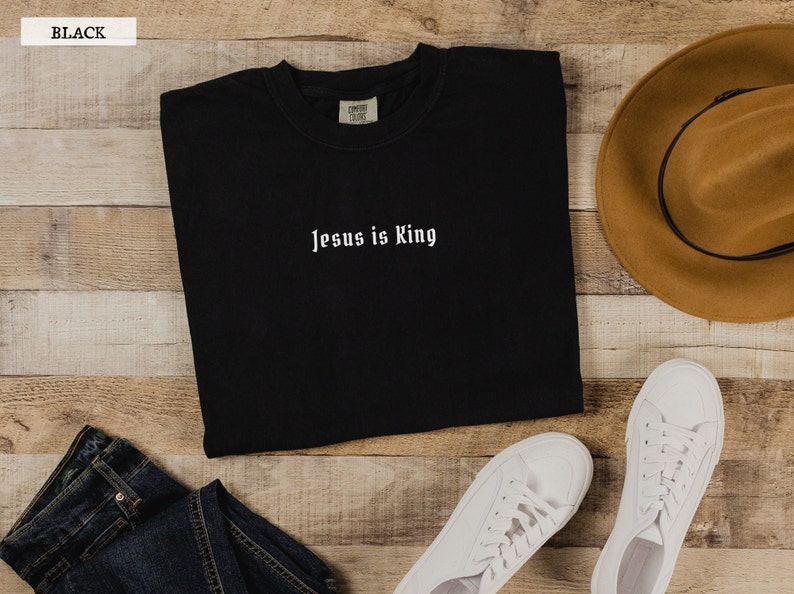 May include: Black long-sleeved t-shirt with the text "Jesus is King" in white lettering. The shirt is laid flat on a wooden surface with a brown hat and white sneakers.