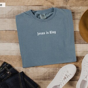 May include: A light blue t-shirt with the text "Jesus is King" in white lettering. The shirt is folded and laid on a wooden surface with a pair of white sneakers, a brown hat, and blue jeans.