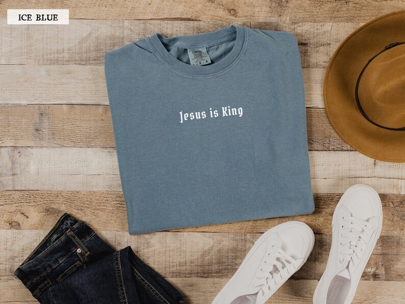 May include: A light blue t-shirt with the text "Jesus is King" in white lettering. The shirt is folded and laid on a wooden surface with a pair of white sneakers, a brown hat, and blue jeans.