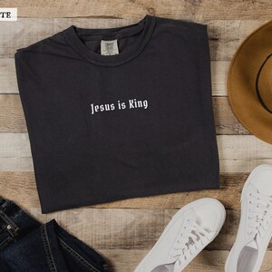 May include: A graphite grey t-shirt with the text "Jesus is King" in white lettering. The shirt is laid flat on a wooden surface with a brown hat, blue jeans, and white sneakers.