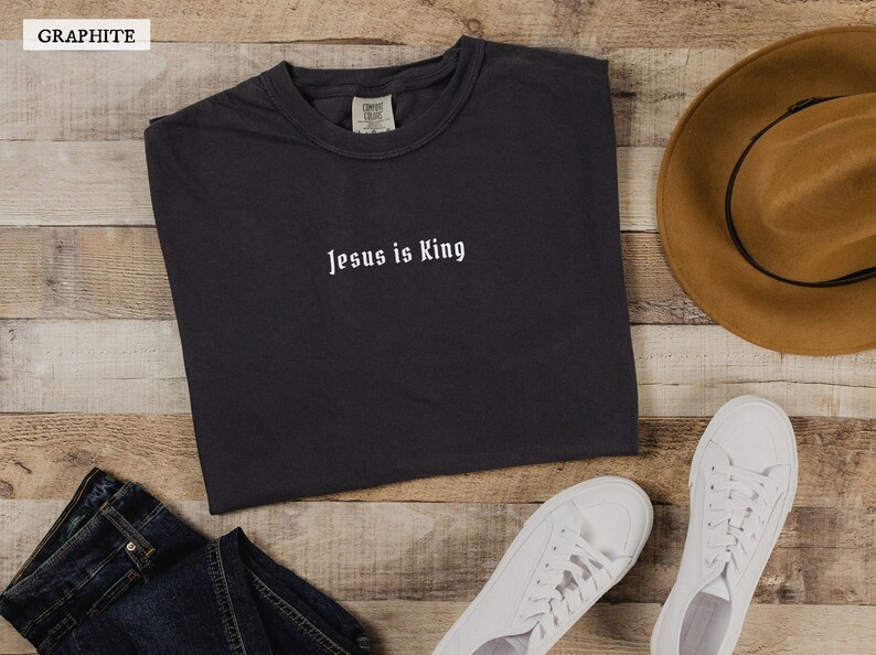 May include: A graphite grey t-shirt with the text "Jesus is King" in white lettering. The shirt is laid flat on a wooden surface with a brown hat, blue jeans, and white sneakers.