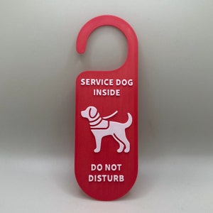 May include: A red door hanger with white text reading "SERVICE DOG INSIDE" and "DO NOT DISTURB". A white graphic of a dog wearing a service vest is below the text. The hanger has a curved top for hanging on a door handle.