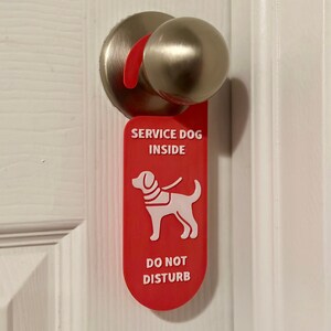 May include: A red door hanger with white text reading "SERVICE DOG INSIDE" and "DO NOT DISTURB". A white graphic of a service dog wearing a vest is also on the hanger. The hanger is on a white door with a silver doorknob.