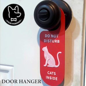 May include: Red door hanger with the words "DO NOT DISTURB" and "CATS INSIDE" in white. A white cat silhouette is featured below the text. The hanger is attached to a black doorknob.