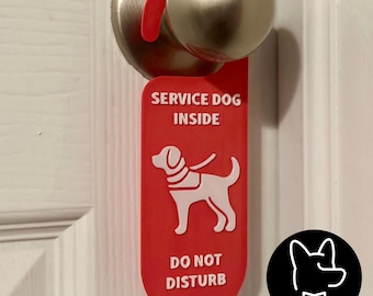 May include: A red door hanger sign with the text "SERVICE DOG INSIDE" and "DO NOT DISTURB" in white. A white graphic of a service dog is also on the sign. The sign is hanging on a door handle.