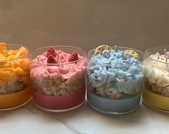 May include: Four dessert-themed candles in clear glass jars. Each candle has a different color and topping, including orange slices, pink frosting with raspberries, blue frosting with waffles, and white frosting with macarons.